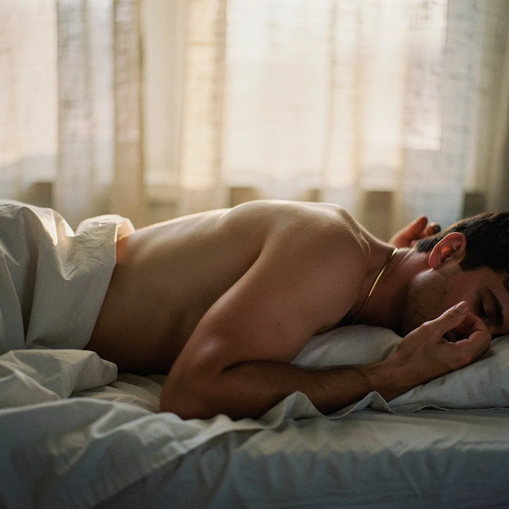 Homme de trente ans aux traits doux allongé sur un lit de lin blanc, lumière tamisée